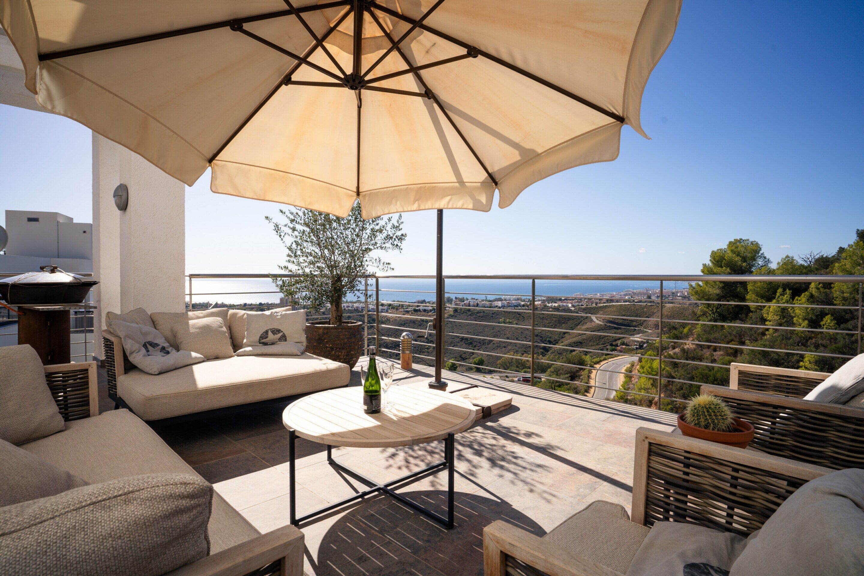 A terrace with a sofa, a table, and an umbrella. There is a view of the landscape and a cactus plant.