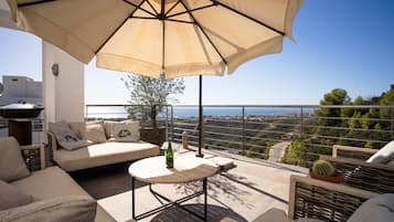 A terrace with a sofa, a table, and an umbrella. There is a view of the landscape and a cactus plant.