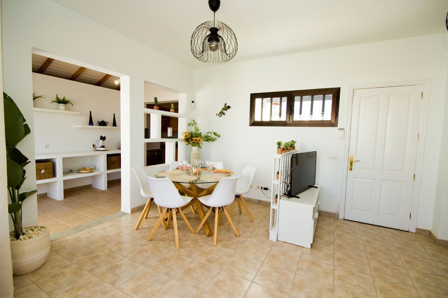 A dining area with a round table, white chairs, and a TV on a stand. There are shelves with plants and a ceiling fan.