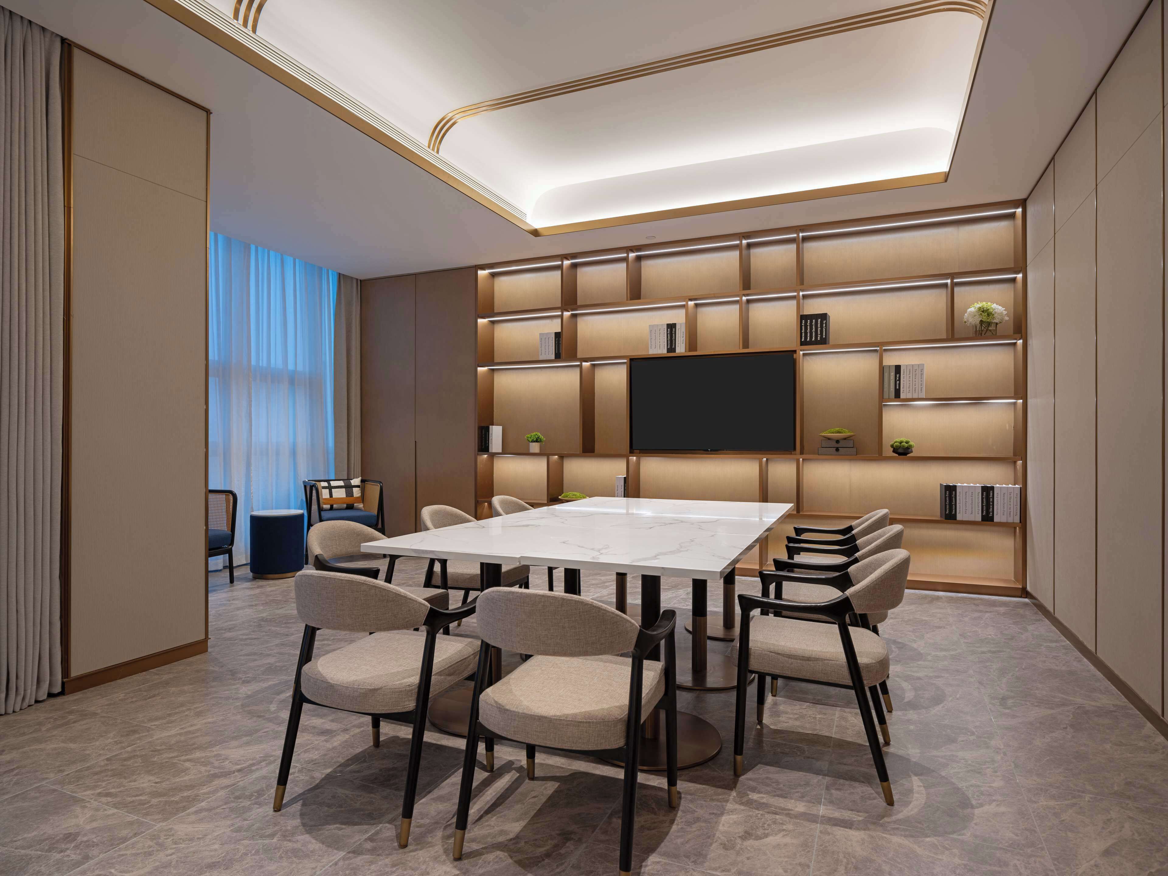Un salón de conferencias moderno con una mesa grande, sillas cómodas, un televisor de pantalla plana y estanterías empotradas con libros y objetos decorativos.