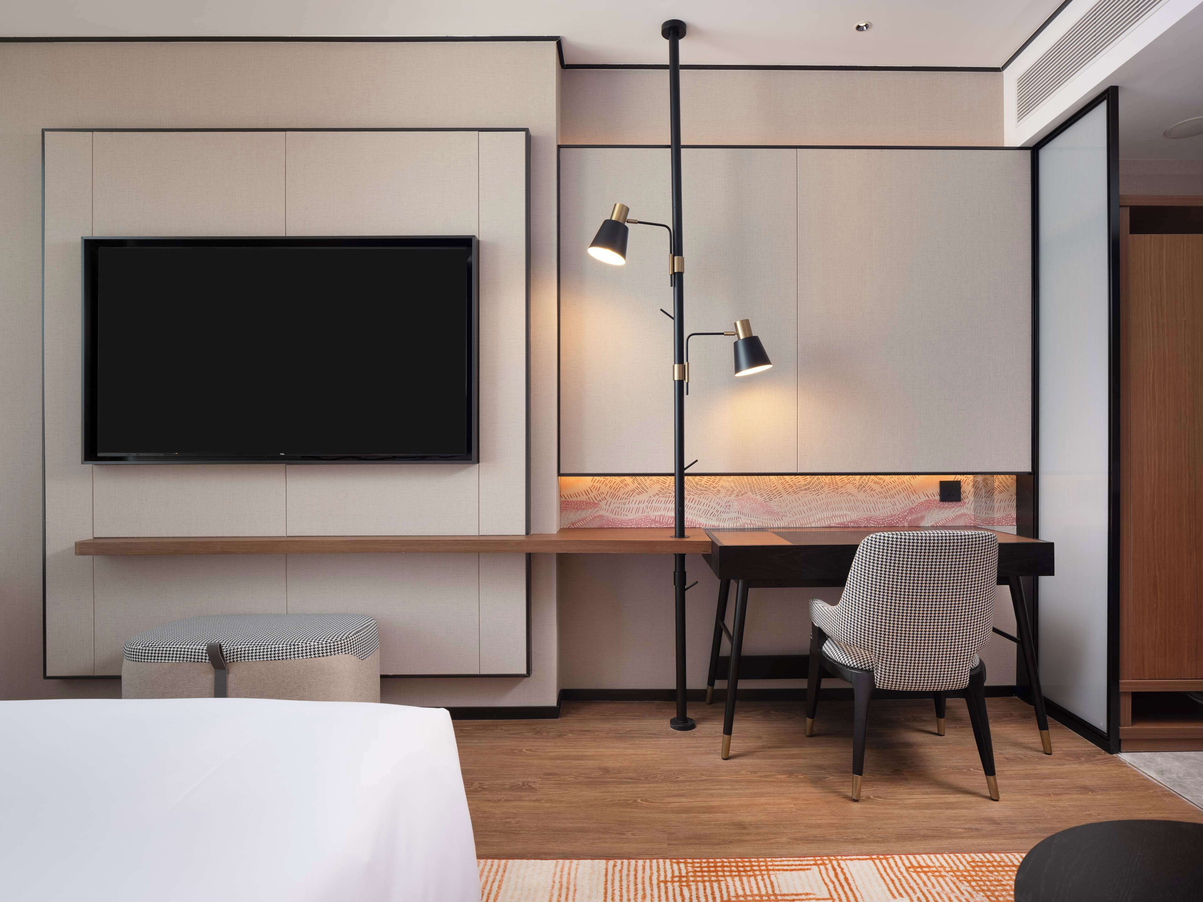 Ein modernes Hotelzimmer mit großem Flachbildfernseher an der Wand, einem Schreibtisch mit Stuhl, einer Stehlampe und einem kleinen Hocker.