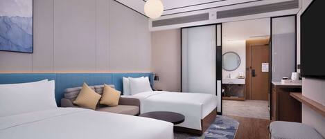 A modern hotel room with two beds, a sofa, a small table, a TV, and a bathroom visible through a sliding door.