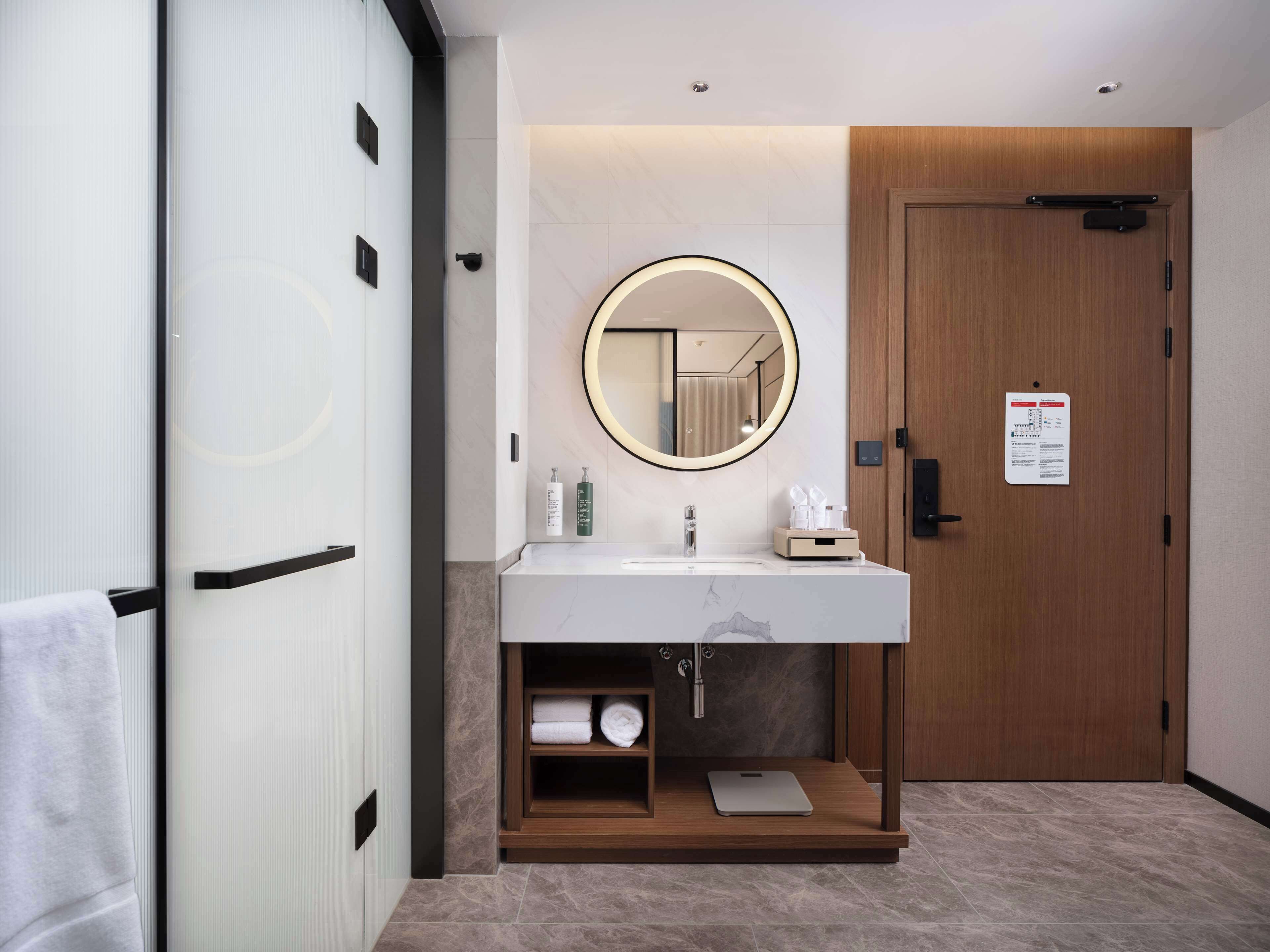 A modern bathroom with a wooden door, a round mirror, a white sink, and a towel rack.