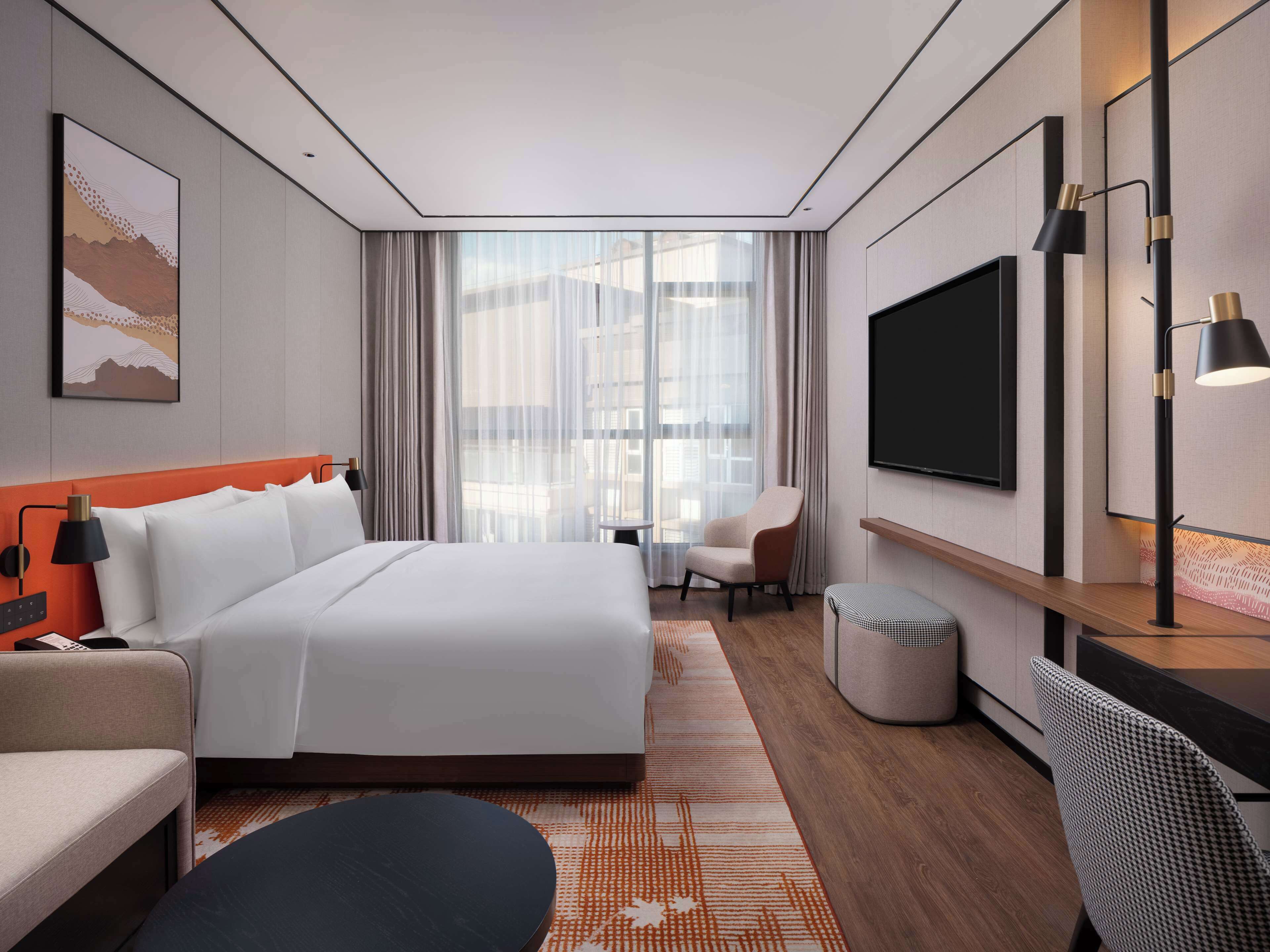 Una habitación de hotel moderna con una cama grande, un escritorio, una silla, un televisor y una ventana con cortinas.