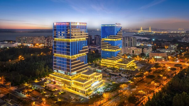 A high-rise hotel with a prominent Hilton Garden Inn sign, surrounded by urban infrastructure and a body of water in the background. - Hilton Garden Inn Danzhou Yangpu (Danzhou)