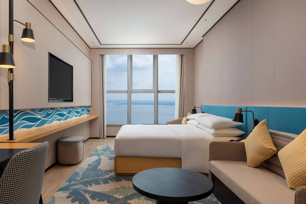 A modern hotel room with a large window, a bed, a sofa, a small table, and a TV on the wall.