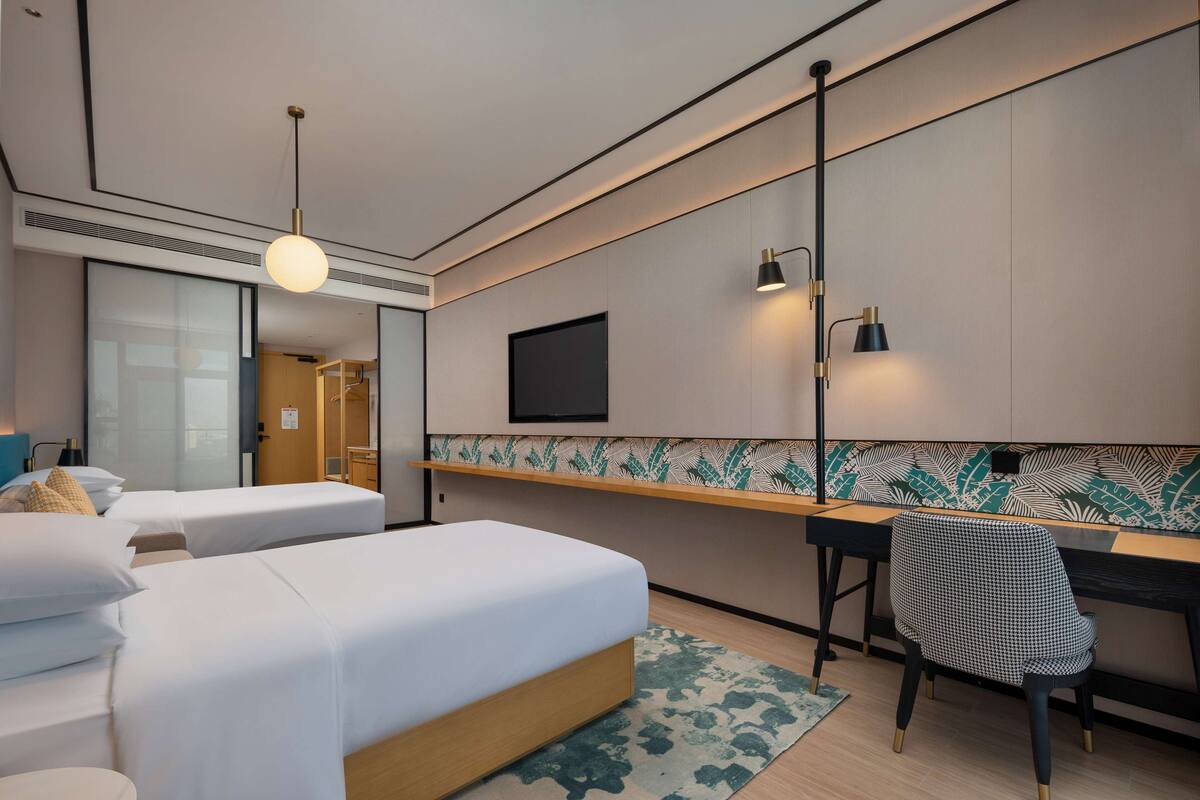 A hotel room with two beds, a desk, a chair, a TV, and a patterned wall.