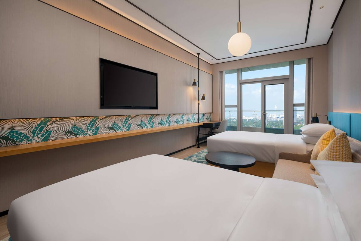 A hotel room with two beds, a TV mounted on the wall, a large window with a city view, and a round table.