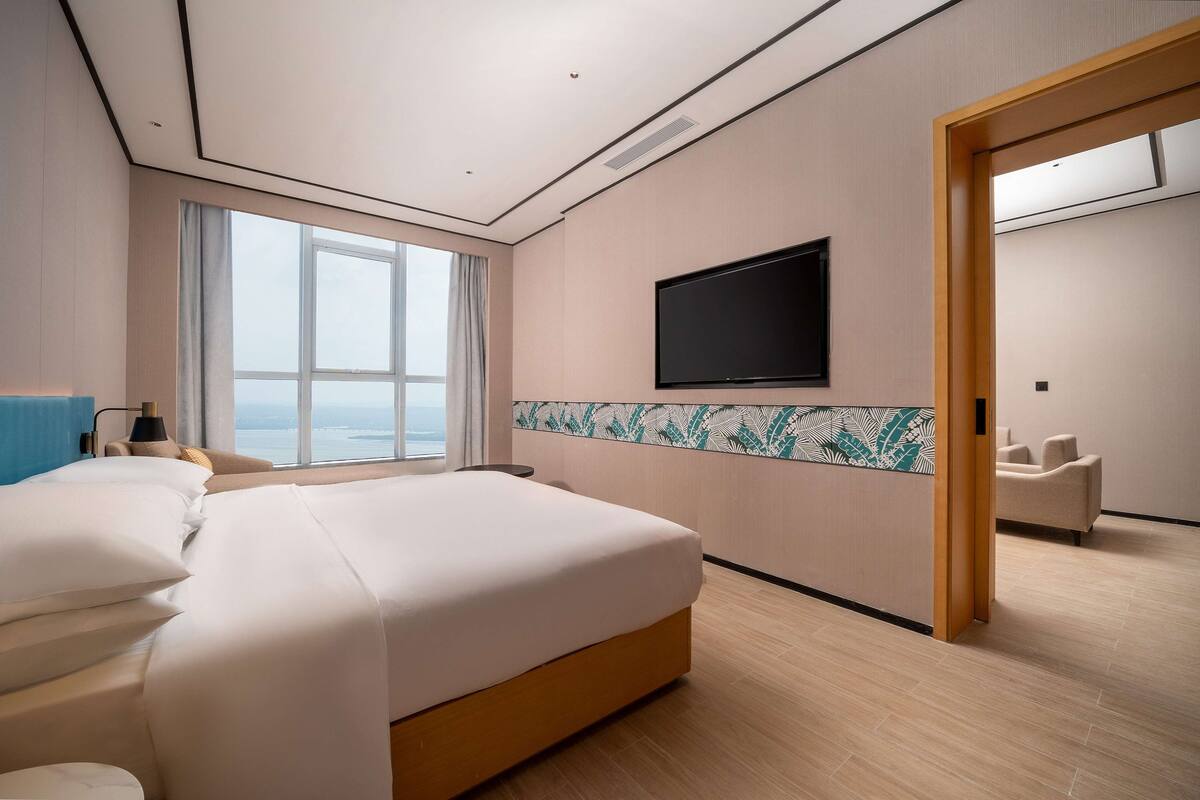 A hotel room with a large bed, a TV mounted on the wall, and a view of the sea through the window.