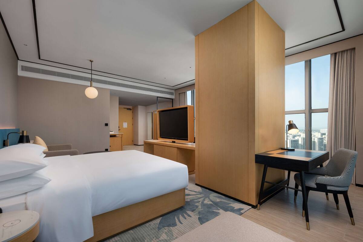 A hotel room with a large bed, a desk, a chair, a TV, and a window with a city view.