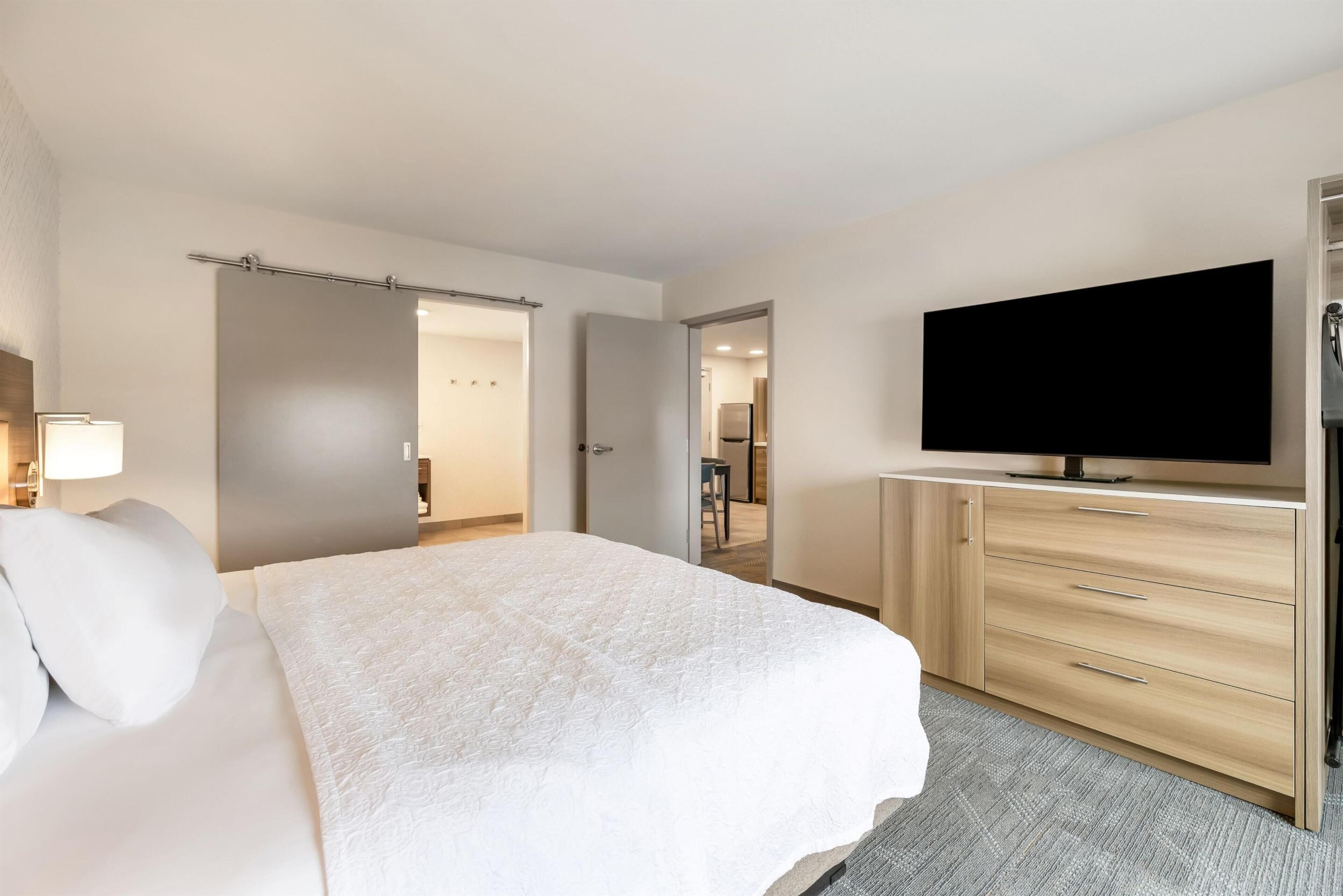 A hotel room with a bed, a TV, a wooden dresser, and a door leading to a bathroom.