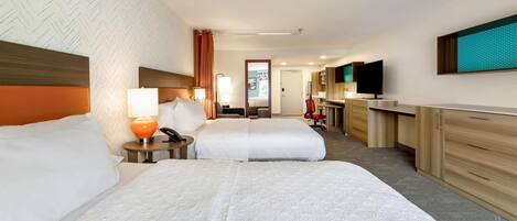 Quarto de hotel com duas camas, uma mesa de trabalho, televisão e banheiro ao fundo.