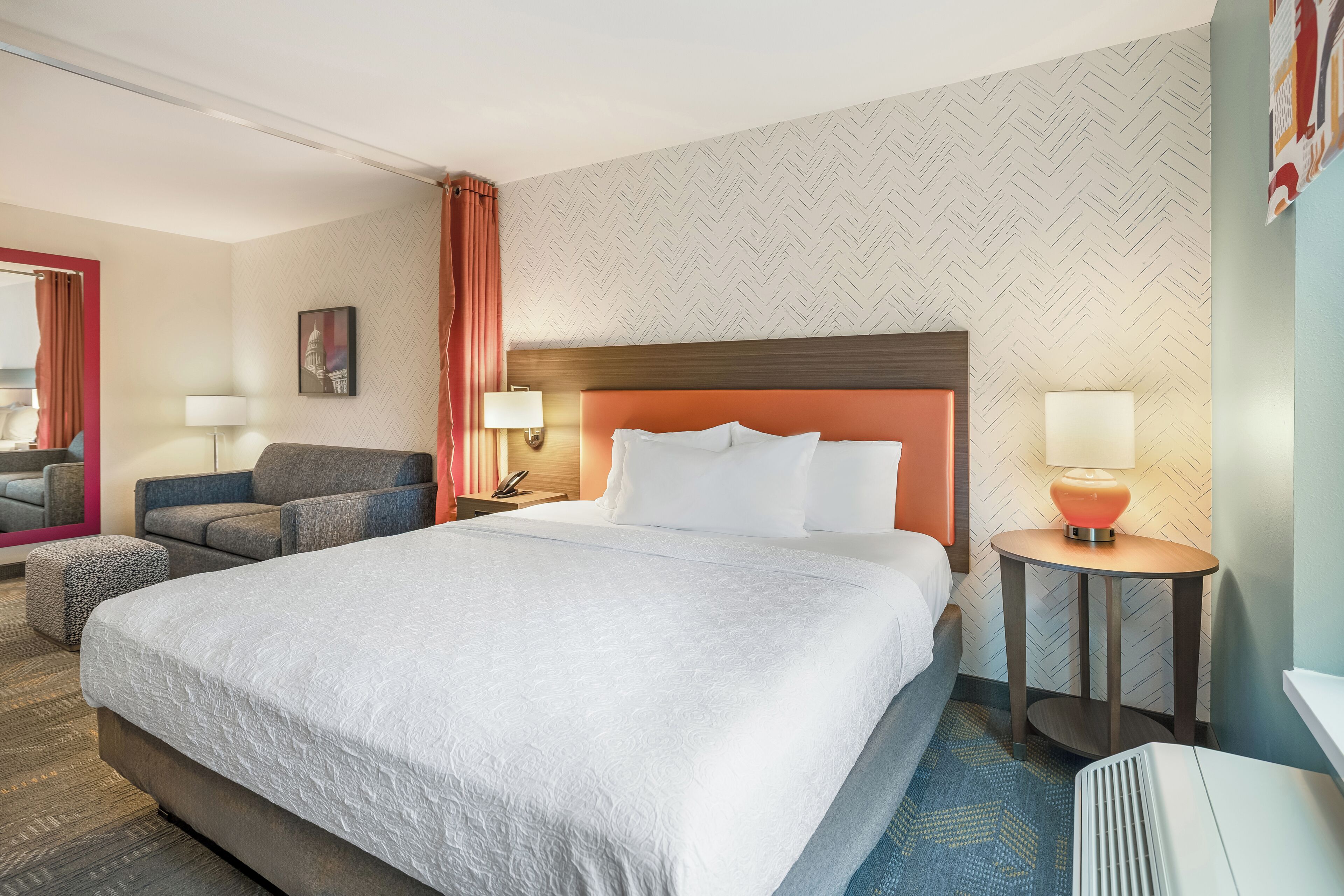 Una habitación de hotel con una cama grande, un sofá, una mesita de noche con lámpara, un espejo y un papel tapiz estampado.