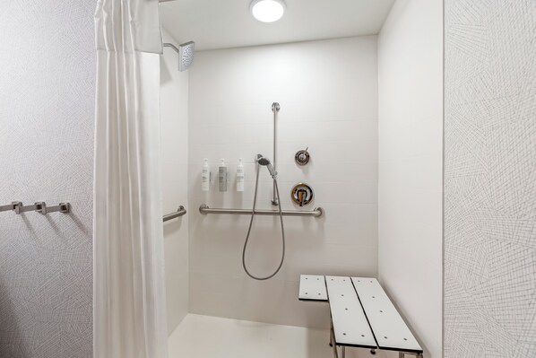 A white bathroom with a shower, a towel rack, and a bench. - Home2 Suites By Hilton Madison East (Madison)