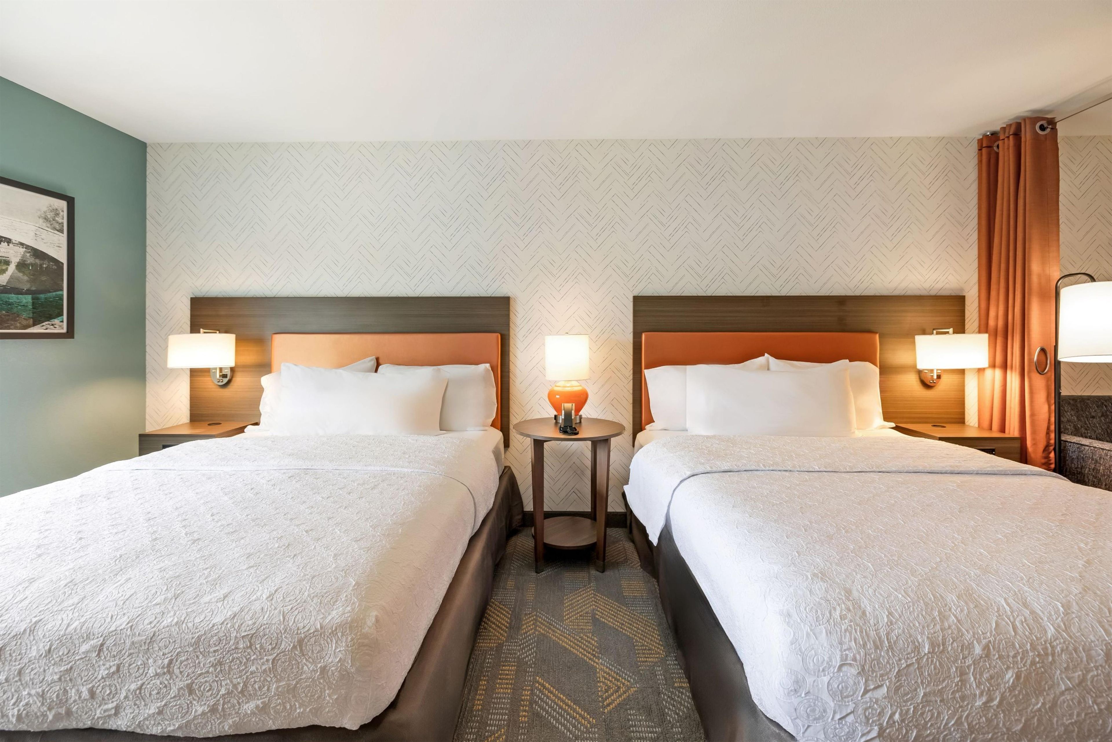 A hotel room with two beds, a small table, and a wall-mounted lamp.