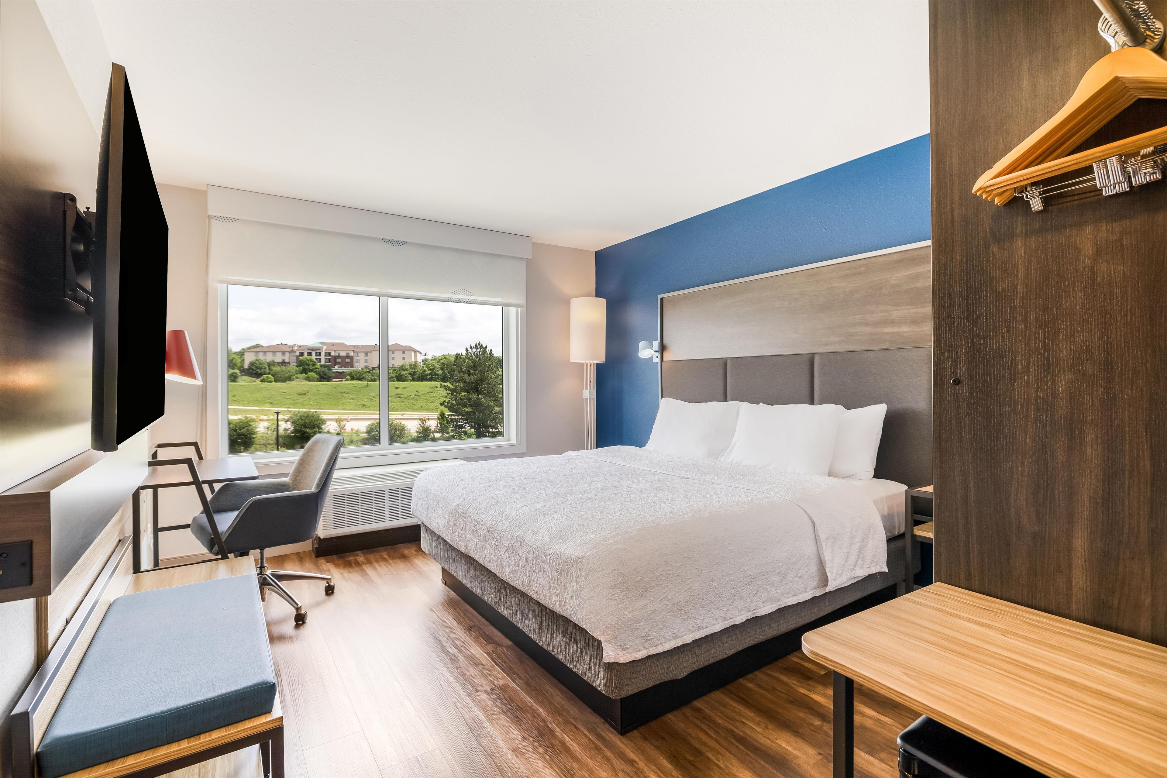 A modern hotel room with a large bed, a desk, a chair, a TV, and a window with a view of greenery and buildings.