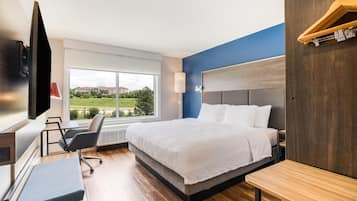 A modern hotel room with a large bed, a desk, a chair, a TV, and a window with a view of greenery and buildings.