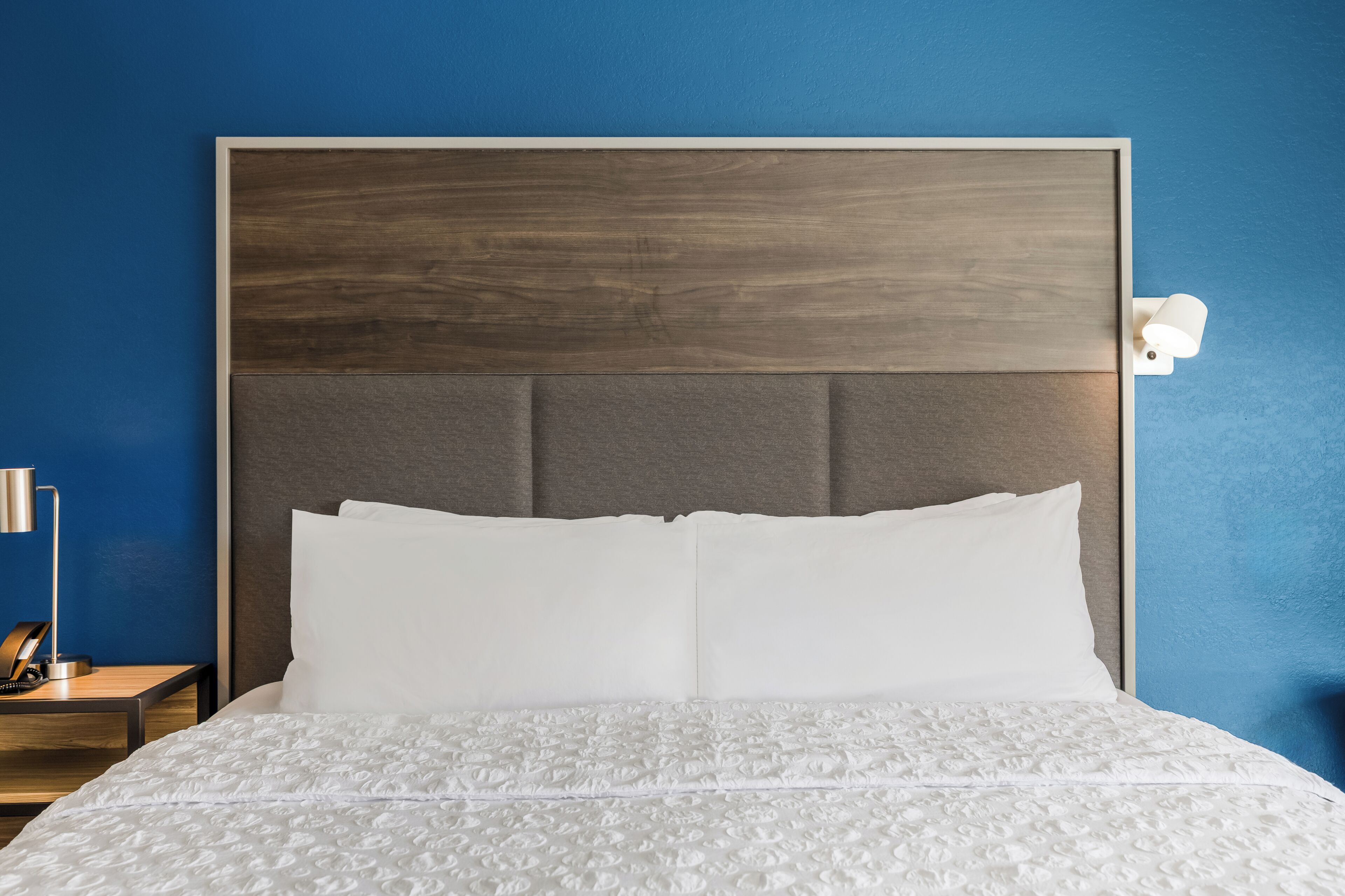 A bed with a headboard, white bedding, and a nightstand with a lamp and phone.