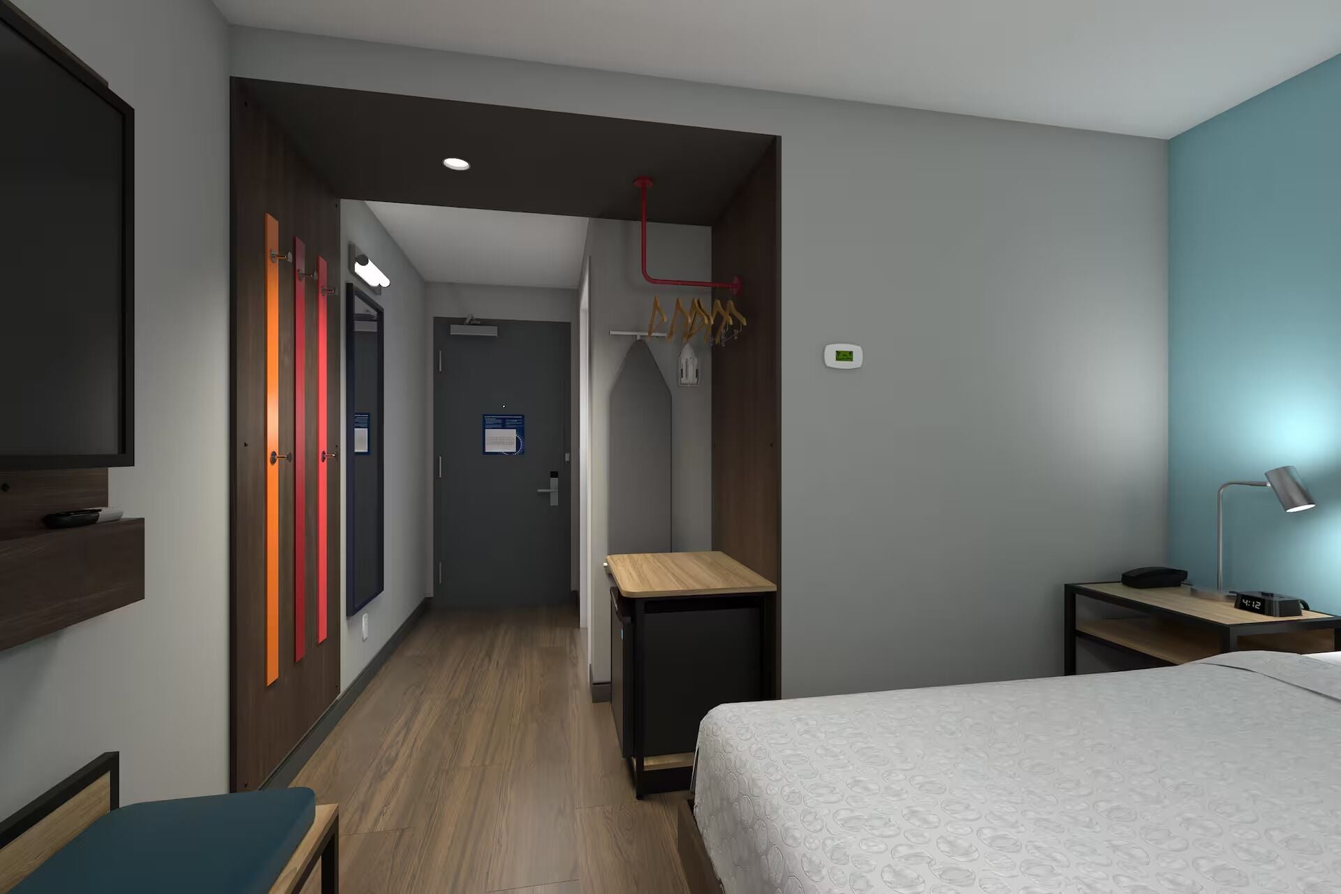 A hotel room with a bed, bedside table, lamp, TV, and a hallway leading to a bathroom.