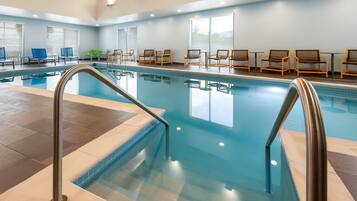 Ein heller, überdachter Pool mit klarem Wasser, umgeben von Liegestühlen und Tischen, große Fenster sorgen für Tageslicht.