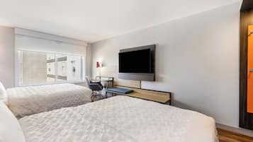 Ein Hotelzimmer mit zwei Betten, einem Schreibtisch, einem Stuhl, einem Fernseher und einem Fenster mit Blick auf Gebäude.