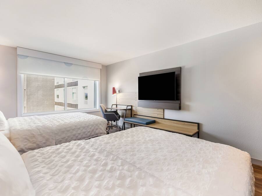 A hotel room with two beds, a desk, a chair, a television, and a window with a view of buildings.