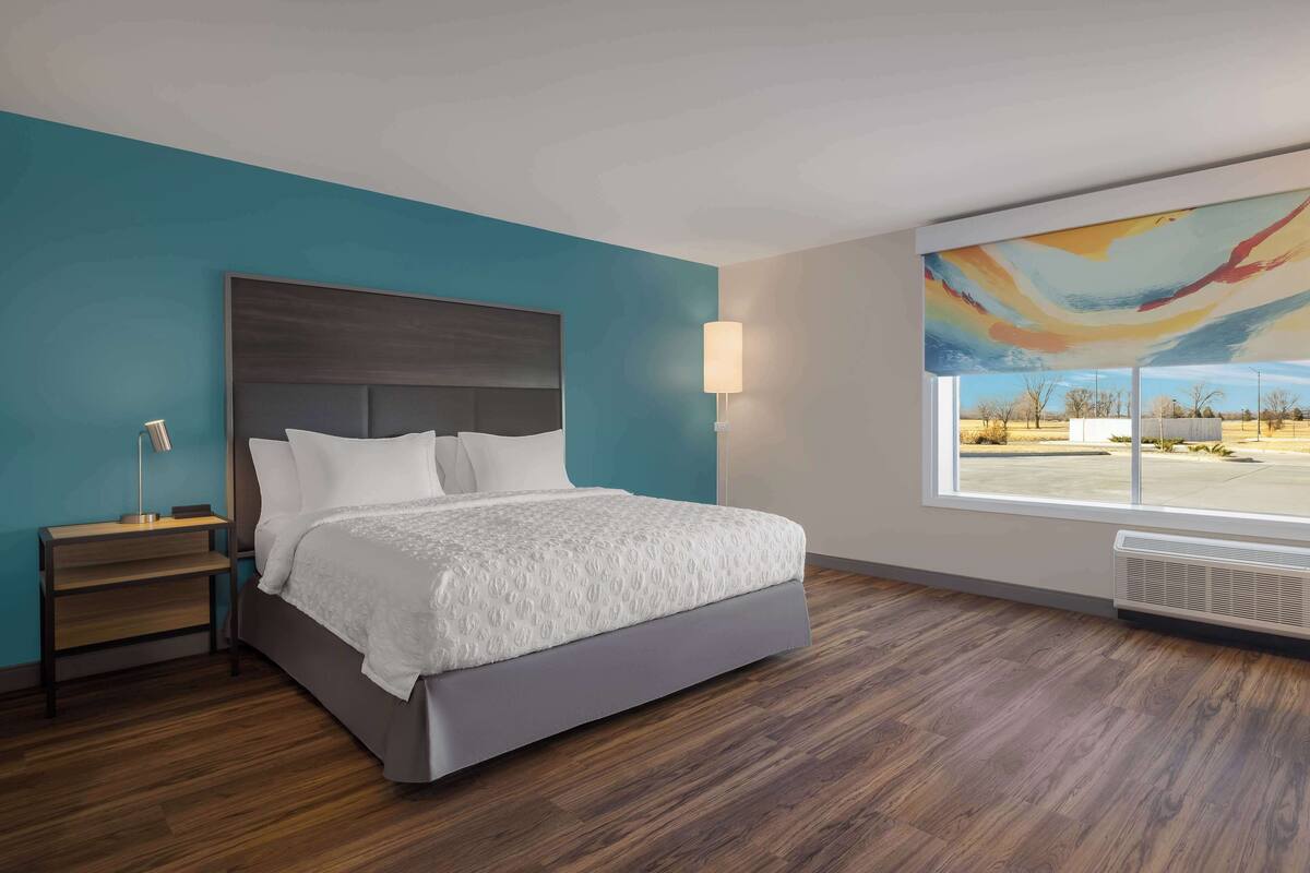 Una habitación de hotel moderna con una cama grande, una mesita de noche, una lámpara y vista a un estacionamiento a través de un ventanal amplio.