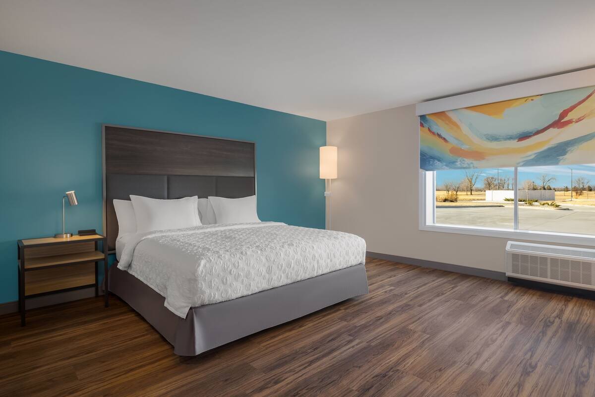Habitación de hotel moderna con una cama grande, una mesita de noche, una lámpara y vistas al exterior a través de un ventanal amplio.