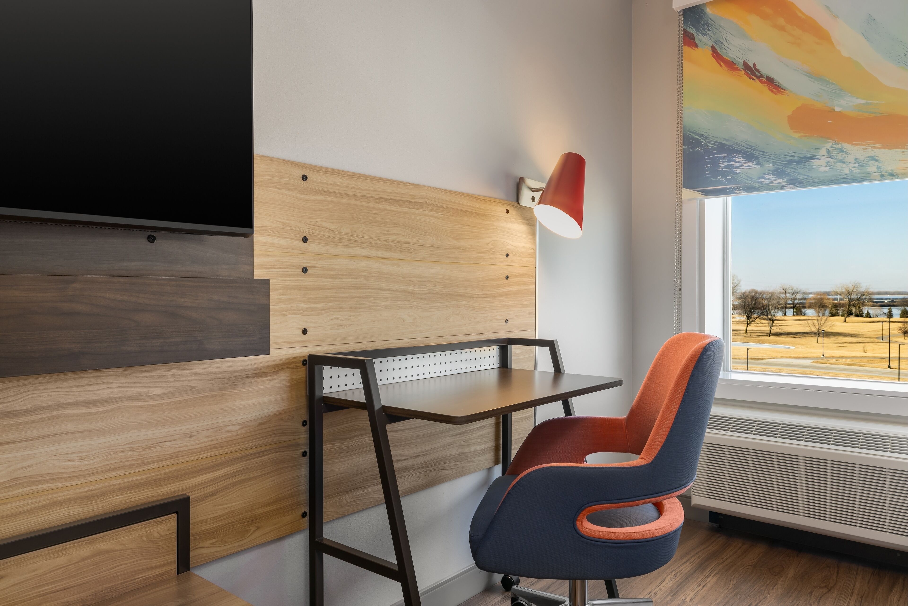 A modern room with a wooden desk, a chair, a TV mounted on the wall, and a window with a view of the outdoors.