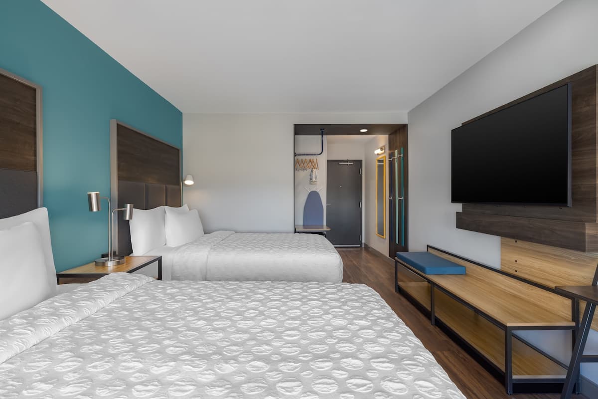 Habitación de hotel con dos camas, un escritorio, un televisor y un baño visible a través de una puerta abierta.