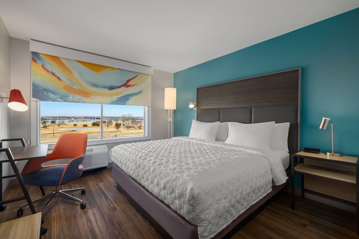 Una habitación de hotel moderna con una cama grande, un escritorio, una silla, vistas al exterior y un cuadro abstracto y colorido en la pared.