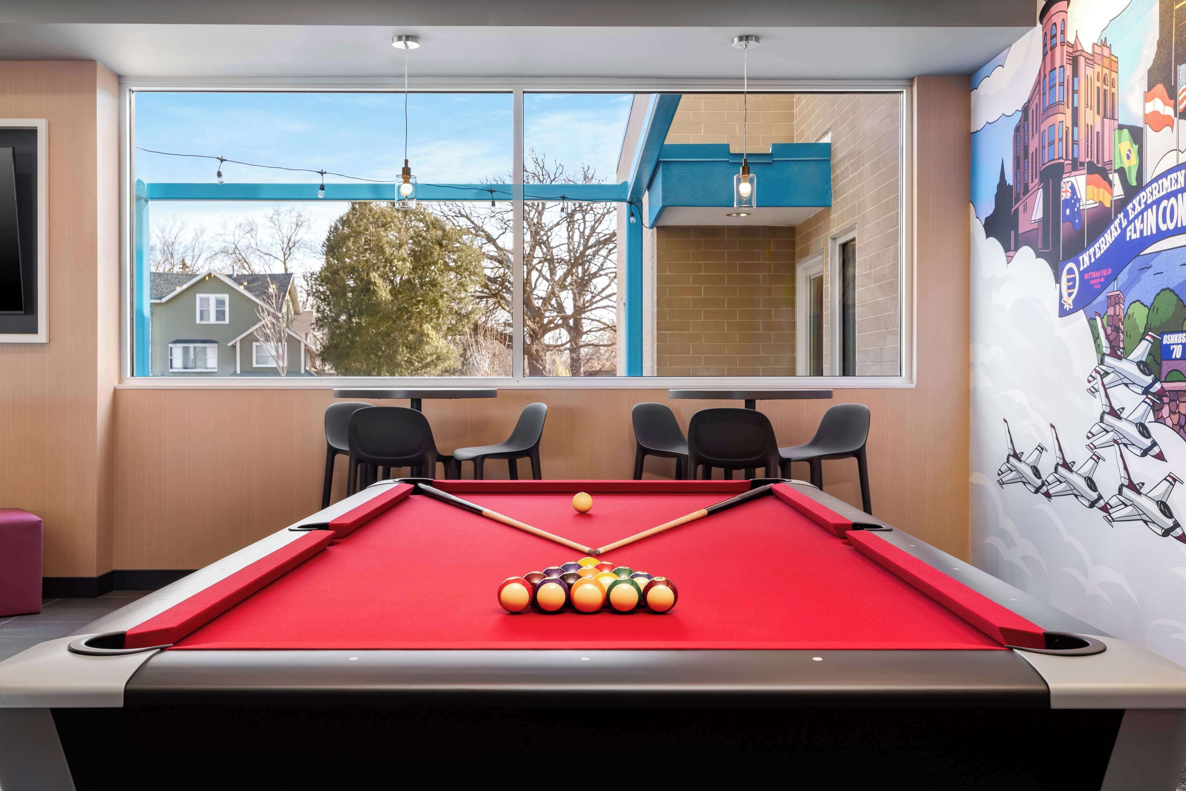 A pool table with a red felt surface, a cue stick, and a set of billiard balls. There are two chairs at the table, and a mural depicting an airplane and a building on the wall.