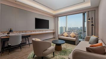 Una habitación de hotel moderna con una televisión de pantalla plana montada en la pared, un sofá, un escritorio con una silla y un ventanal con vistas a la ciudad.