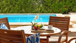 Une table ronde en bois, sur laquelle se trouvent un vase blanc avec des fleurs, un panier de fruits, ainsi qu’une tasse et une soucoupe. Deux chaises en bois, dotées de coussins bleus et blancs, sont placées autour de la table. On aperçoit une piscine à l’arrière-plan.