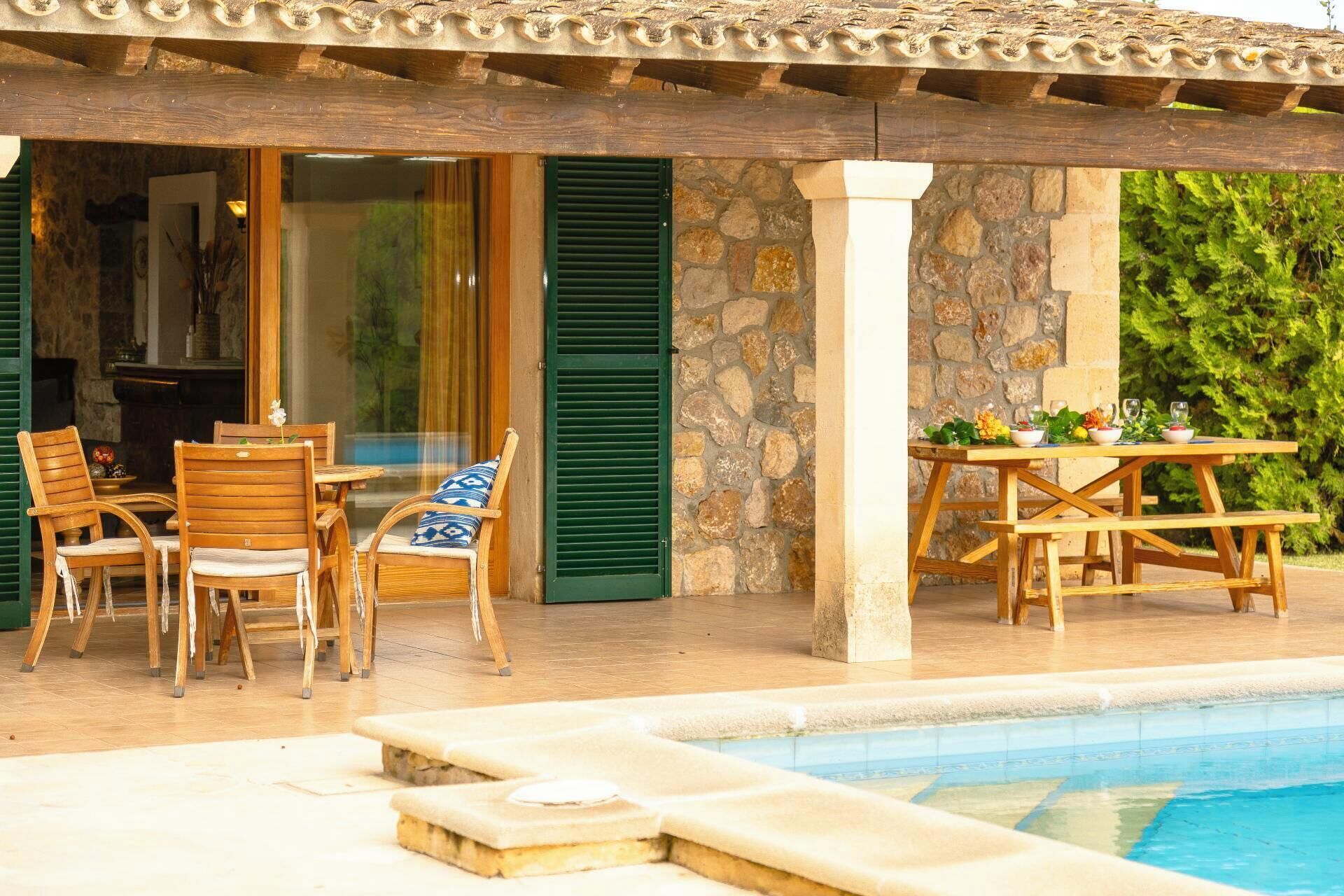 Zona junto a la piscina con mobiliario de madera, pared de piedra y vista a un paisaje verde exuberante.