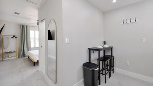 Een moderne, goed verlichte kamer met een bed, een klein tafeltje met stoel, een prullenbak en een kapstok.