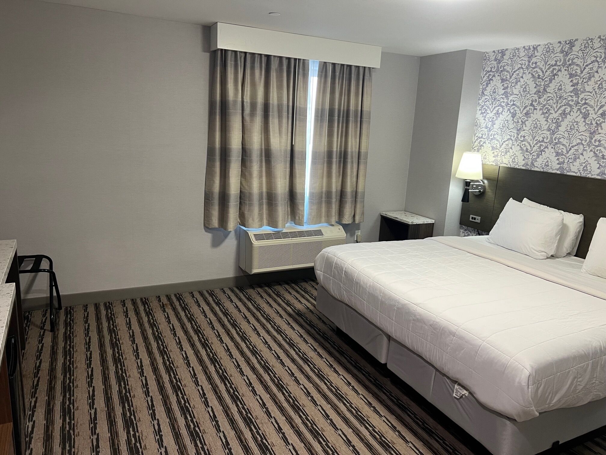 Ein Hotelzimmer mit Bett, Schreibtisch, Stuhl, Nachttisch, Lampe, einem Fenster mit Vorhängen und einem Heizkörper.