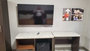 Une pièce dotée d’un grand téléviseur à écran plat fixé au mur, d’un petit réfrigérateur placé sous un bureau et d’une armoire en bois sur le côté gauche.
