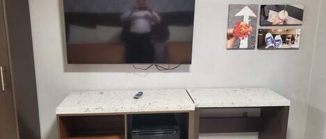 A room with a large flat-screen TV mounted on the wall, a small refrigerator under a desk, and a wooden cabinet to the left.