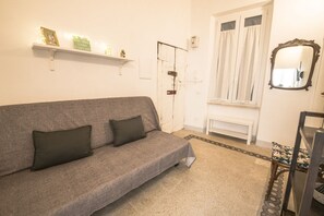 A room with a grey sofa, a white door, a window with curtains, a small white table, and a mirror on the wall.