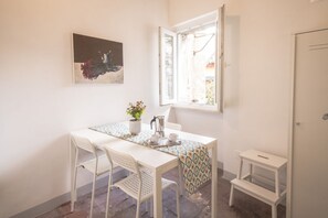 A small dining area with a table, chairs, a window, a painting, and a small white stool.