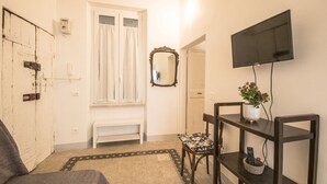 Ein Zimmer mit einem Bett, einem an der Wand befestigten Fernseher, einem Spiegel, einer Bank und einem kleinen Tisch mit Pflanze und Korb.