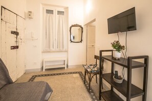 Une pièce comprenant un lit, un téléviseur fixé au mur, un miroir, un banc et une petite table sur laquelle se trouvent une plante et un panier.