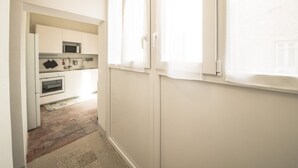 Ein enger Flur führt zu einer Küche mit weißen Schränken, einer Mikrowelle und einem Fenster mit Jalousien.