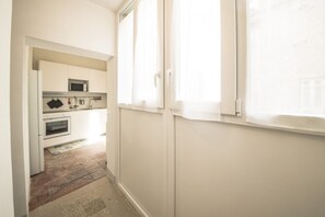 Un couloir étroit mène à une cuisine dotée de meubles blancs, d’un four à micro-ondes et d’une fenêtre avec des stores.