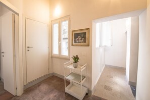 Un couloir avec des portes blanches, une fenêtre, un tableau encadré et une étagère blanche sur laquelle est posée une plante.