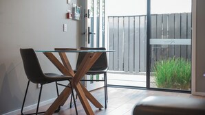 A modern dining area with a glass table and two chairs, a wooden floor, and a sliding glass door leading to a balcony with a wooden fence and greenery.