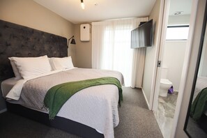 Un lit bien fait, agrémenté d’un plaid vert, un téléviseur à écran plat fixé au mur et une salle de bain visible à travers une porte ouverte.