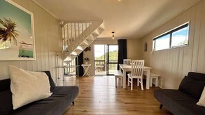 A modern living room with a staircase, wooden flooring, and a dining area with white furniture. - Driftwood Retreat - Tutukaka (Tutukaka)