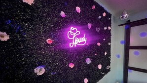 Een kamer met een decoratieve wand waarop een neonbord met de tekst 'Lovely' staat, omringd door bloemen.